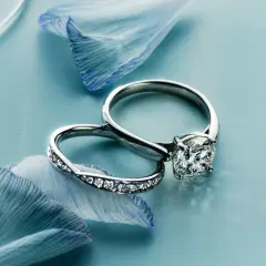 Dos alianzas de boda, una con un gran diamante y otra con pequeños diamantes, sobre pétalos de flor azul.