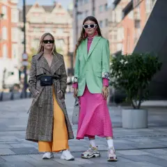 Dos mujeres lucen atuendos vibrantes, mostrando los colores de moda: un abrigo a cuadros con pantalones naranjas y un traje verde menta con vestido rosa fucsia.