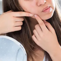 Una joven se mira en un espejo, con los dedos cerca de su mejilla, como si estuviera pensando en cómo explotar un grano.