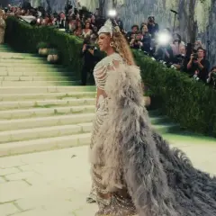 Una mujer desciende escaleras con un vestido de plumas y pedrería, rodeada de fotógrafos. Es un momento icónico de cuando es la Met Gala.