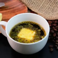 Una taza de café con mantequilla, junto a un trozo de mantequilla y granos de café tostados.