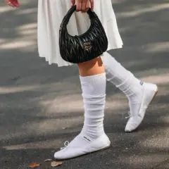 Piernas con bailarinas blancas y calcetines blancos arrugados, sosteniendo un bolso negro.