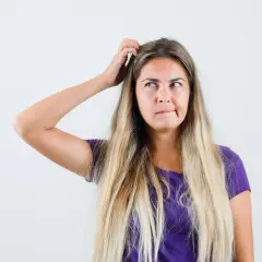 Mujer rubia con el pelo largo y suelto, se rasca la cabeza pensativa, buscando trucos de belleza para un look perfecto.