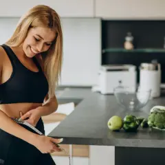 Mujer sonriente se mide la cintura en la cocina, rodeada de alimentos quema grasa por la noche como brócoli y coliflor.