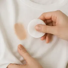 Mano quitando una mancha de maquillaje de una camiseta blanca con un disco de algodón.
