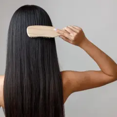 Mujer peinando su largo y oscuro cabello con un cepillo de madera, un gesto clave para saber como nutrir el pelo y mantenerlo sano y brillante.