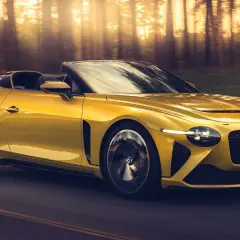 Un Bentley Mulliner Bacalar amarillo brillante recorre un camino boscoso al atardecer, con los rayos del sol filtrándose entre los árboles.