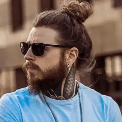 Hombre con barba y pelo recogido en moño, luciendo gafas de sol. Su estilo muestra una transicion pelo corto a largo, con tatuajes visibles en el cuello.