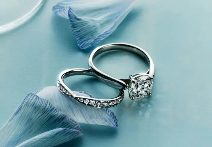 Dos alianzas de boda, una con un gran diamante y otra con pequeños diamantes, sobre pétalos de flor azul.