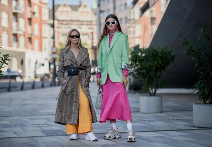 Dos mujeres lucen atuendos vibrantes, mostrando los colores de moda: un abrigo a cuadros con pantalones naranjas y un traje verde menta con vestido rosa fucsia.