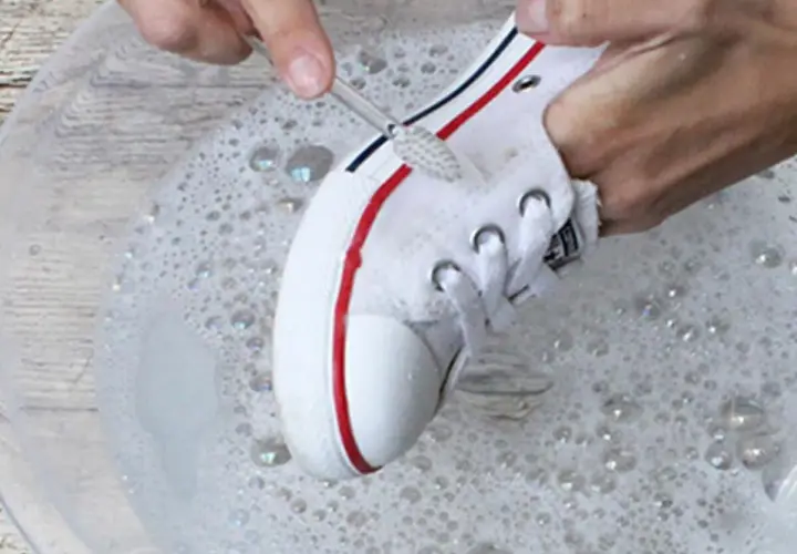 Manos limpian zapatillas blancas de tela con un cepillo en agua con jabón.