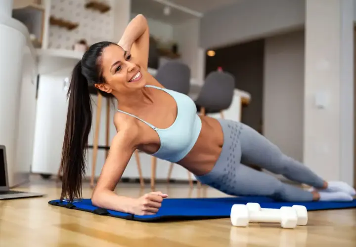 Mujer sonriente realiza una rutina de ejercicios en casa, haciendo plancha lateral sobre una esterilla azul. A su lado, pesas y un portátil.