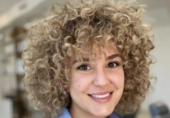 Sonrisa radiante de una mujer con un voluminoso flequillo pelo rizado rubio.