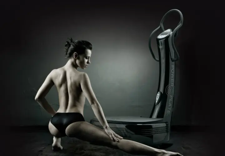 Mujer en pose de estiramiento junto a una plataforma vibratoria, destacando los plataforma vibratoria beneficios para el cuerpo.