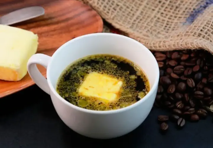 Una taza de café con mantequilla, junto a un trozo de mantequilla y granos de café tostados.
