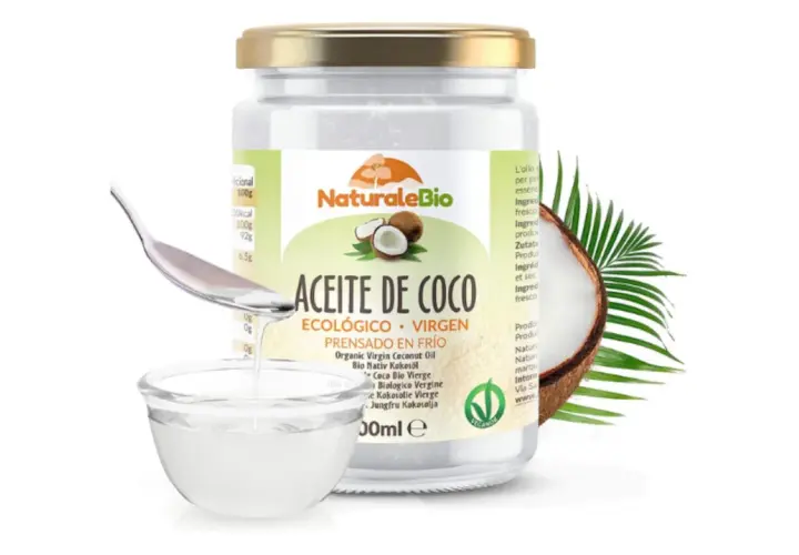 Un frasco de aceite de coco NaturaleBio con una cuchara vertiendo aceite en un cuenco. El aceite de coco es bueno para el pelo, nutriéndolo y dándole brillo.