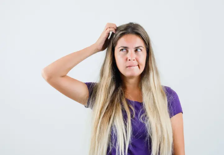 Mujer rubia con el pelo largo y suelto, se rasca la cabeza pensativa, buscando trucos de belleza para un look perfecto.