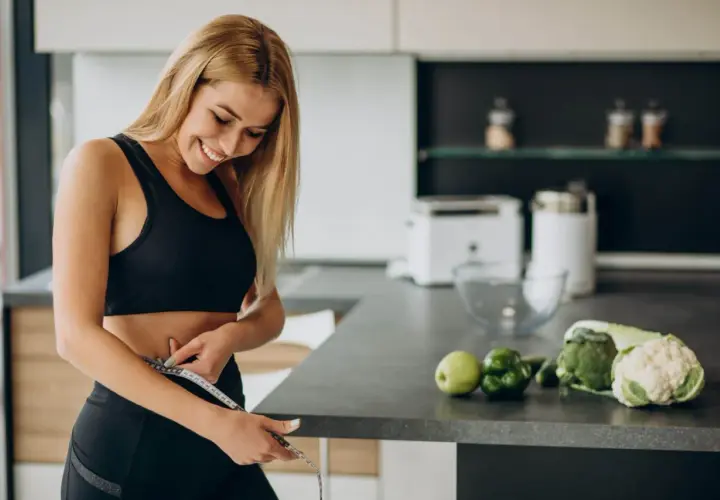 Mujer sonriente se mide la cintura en la cocina, rodeada de alimentos quema grasa por la noche como brócoli y coliflor.