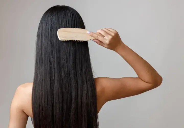 Mujer peinando su largo y oscuro cabello con un cepillo de madera, un gesto clave para saber como nutrir el pelo y mantenerlo sano y brillante.