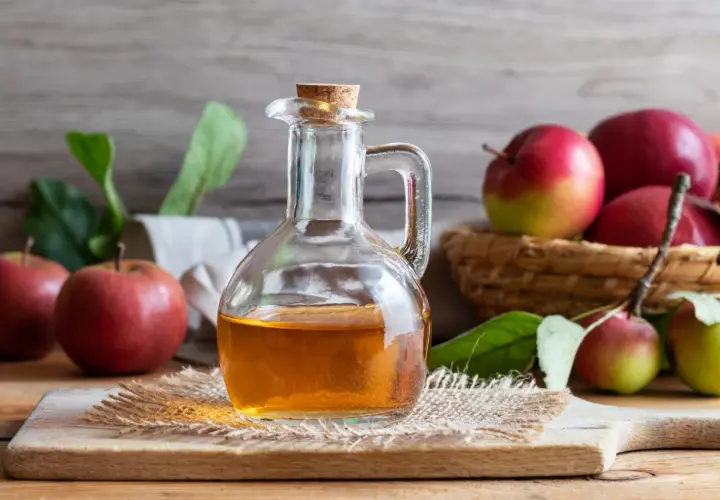 Botella de vidrio con vinagre de sidra de manzana, rodeada de manzanas rojas y verdes frescas.