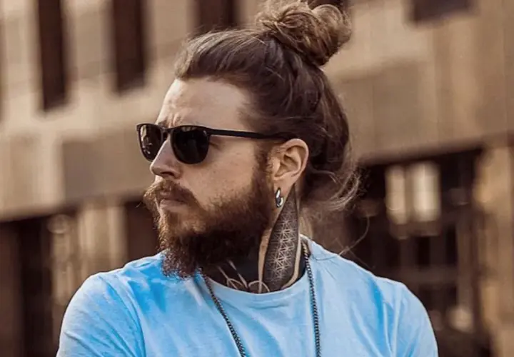 Hombre con barba y pelo recogido en moño, luciendo gafas de sol. Su estilo muestra una transicion pelo corto a largo, con tatuajes visibles en el cuello.