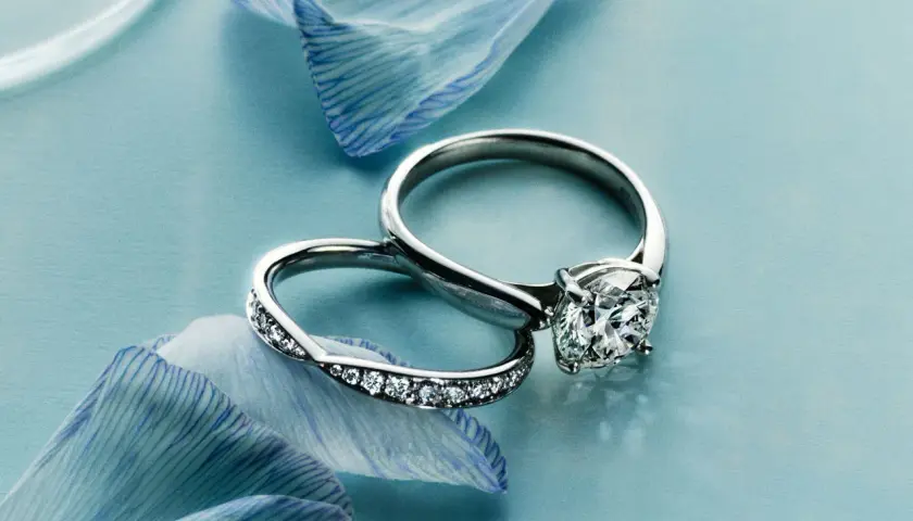 Dos alianzas de boda, una con un gran diamante y otra con pequeños diamantes, sobre pétalos de flor azul.