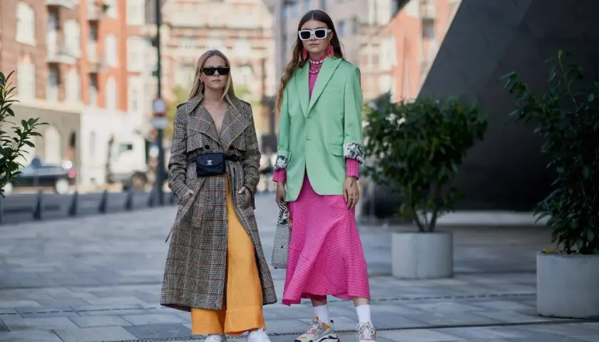 Dos mujeres lucen atuendos vibrantes, mostrando los colores de moda: un abrigo a cuadros con pantalones naranjas y un traje verde menta con vestido rosa fucsia.
