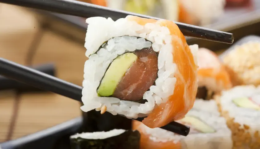 Palillos sostienen un rollo de sushi con salmón y aguacate. ¡Cuidado, el sushi engorda si no te controlas!