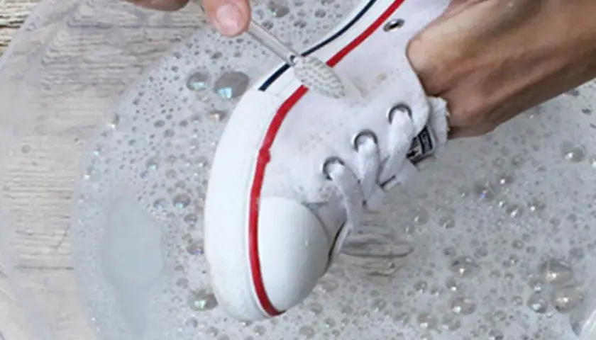 Manos limpian zapatillas blancas de tela con un cepillo en agua con jabón.