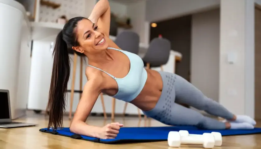 Mujer sonriente realiza una rutina de ejercicios en casa, haciendo plancha lateral sobre una esterilla azul. A su lado, pesas y un portátil.