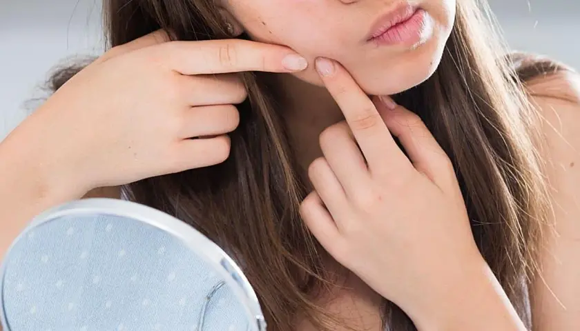 Una joven se mira en un espejo, con los dedos cerca de su mejilla, como si estuviera pensando en cómo explotar un grano.