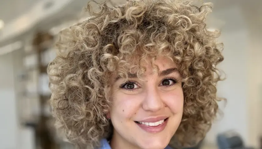 Sonrisa radiante de una mujer con un voluminoso flequillo pelo rizado rubio.