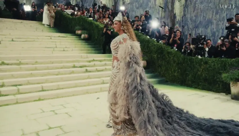 Una mujer desciende escaleras con un vestido de plumas y pedrería, rodeada de fotógrafos. Es un momento icónico de cuando es la Met Gala.