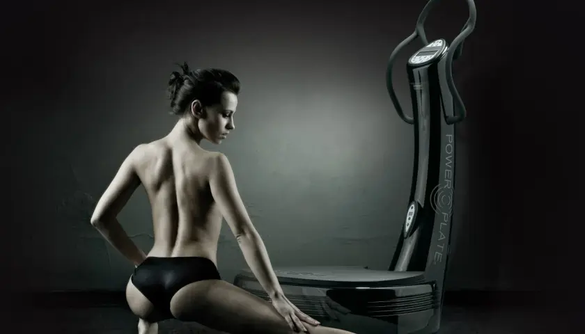 Mujer en pose de estiramiento junto a una plataforma vibratoria, destacando los plataforma vibratoria beneficios para el cuerpo.