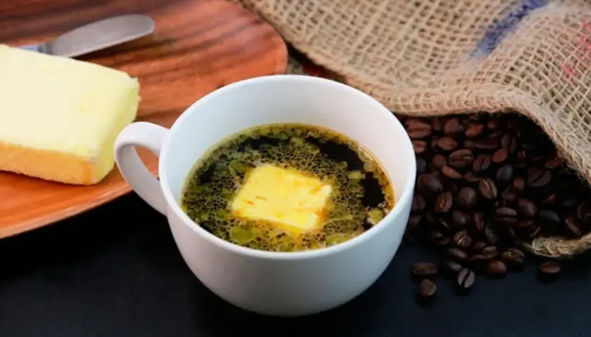 Una taza de café con mantequilla, junto a un trozo de mantequilla y granos de café tostados.