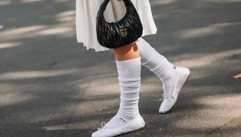 Piernas con bailarinas blancas y calcetines blancos arrugados, sosteniendo un bolso negro.
