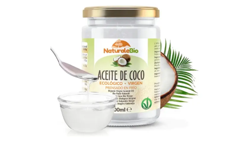 Un frasco de aceite de coco NaturaleBio con una cuchara vertiendo aceite en un cuenco. El aceite de coco es bueno para el pelo, nutriéndolo y dándole brillo.