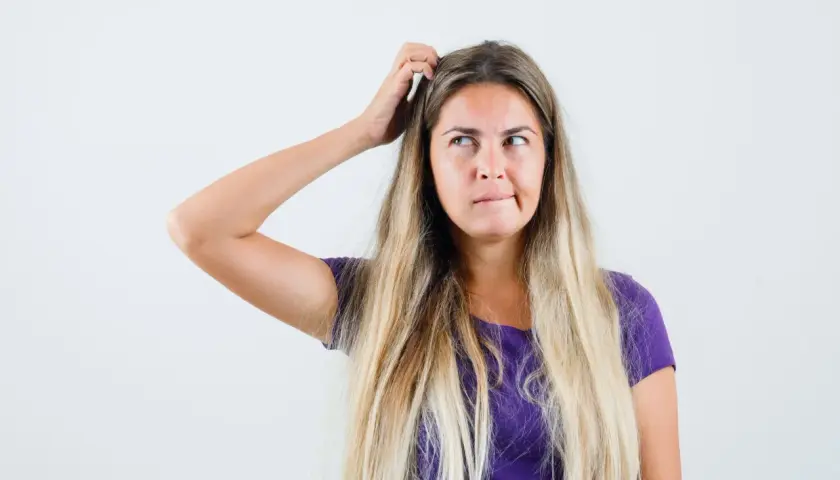 Mujer rubia con el pelo largo y suelto, se rasca la cabeza pensativa, buscando trucos de belleza para un look perfecto.