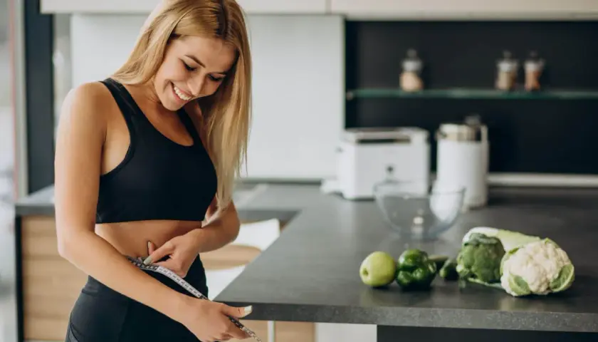 Mujer sonriente se mide la cintura en la cocina, rodeada de alimentos quema grasa por la noche como brócoli y coliflor.