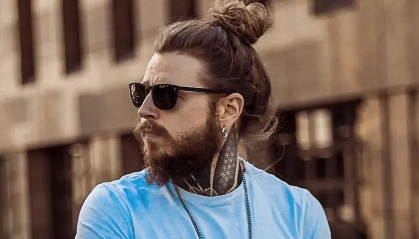 Hombre con barba y pelo recogido en moño, luciendo gafas de sol. Su estilo muestra una transicion pelo corto a largo, con tatuajes visibles en el cuello.