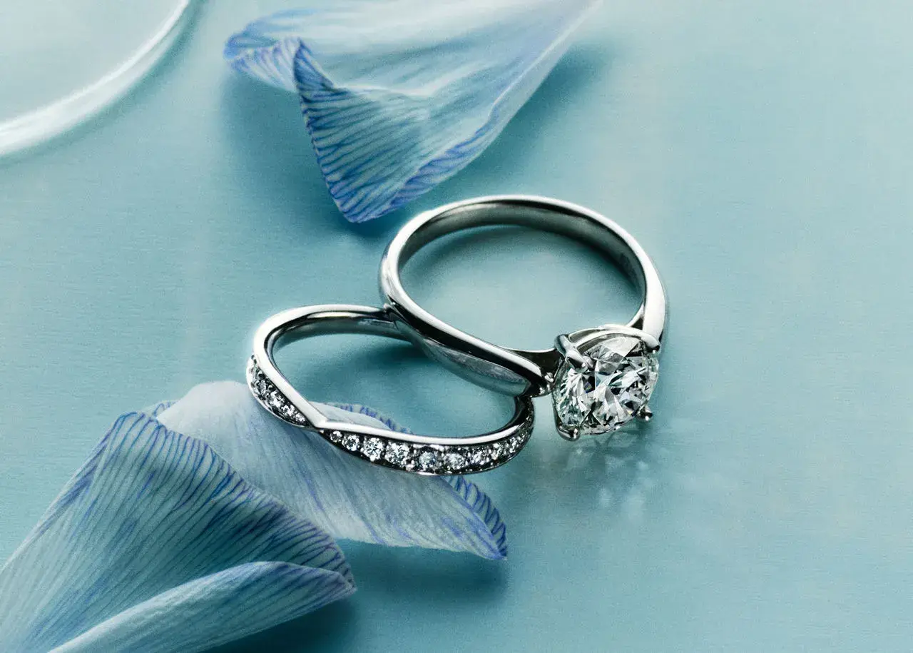 Dos alianzas de boda, una con un gran diamante y otra con pequeños diamantes, sobre pétalos de flor azul.
