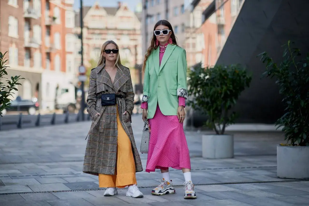 Dos mujeres lucen atuendos vibrantes, mostrando los colores de moda: un abrigo a cuadros con pantalones naranjas y un traje verde menta con vestido rosa fucsia.