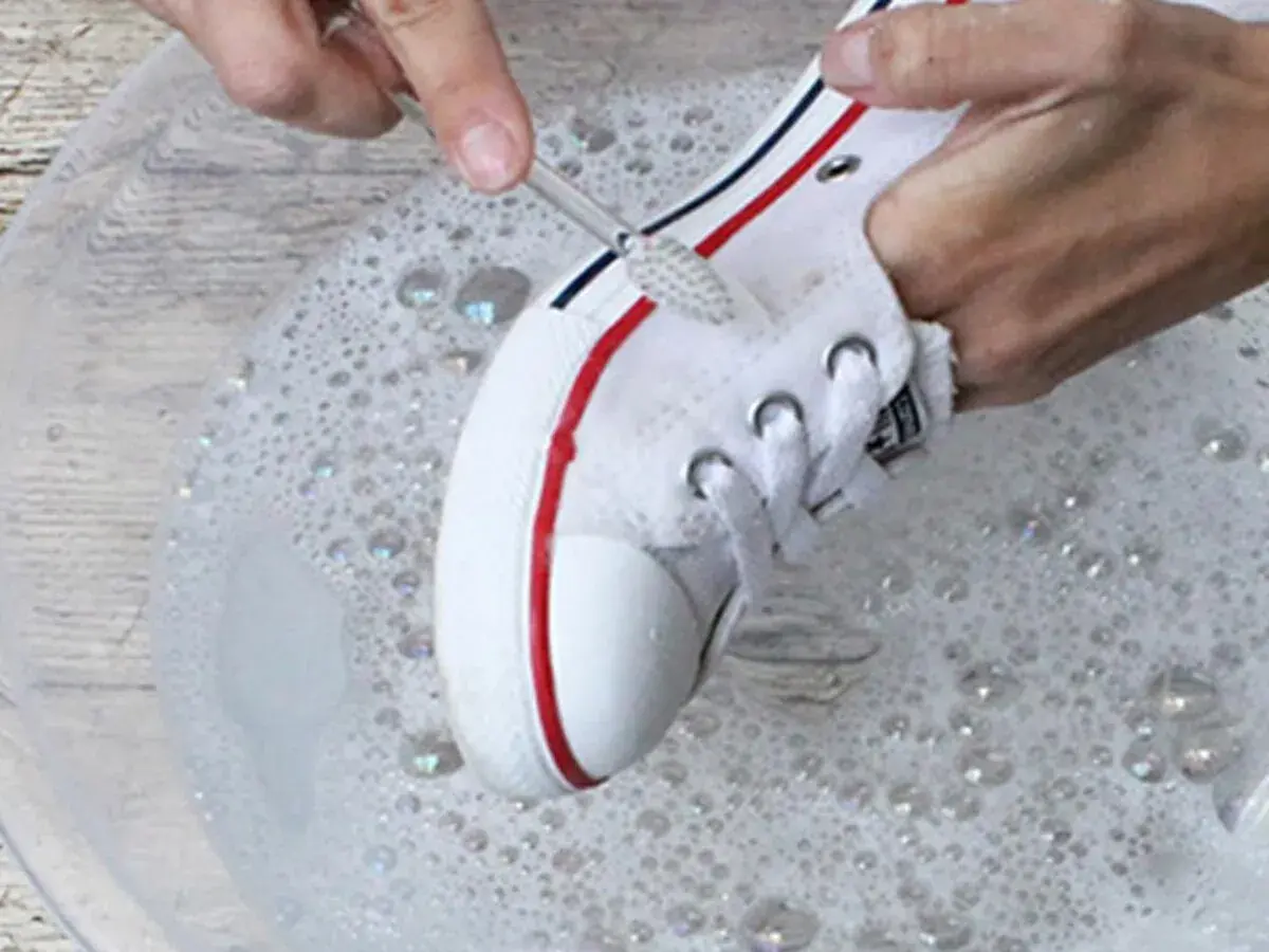 Manos limpian zapatillas blancas de tela con un cepillo en agua con jabón.