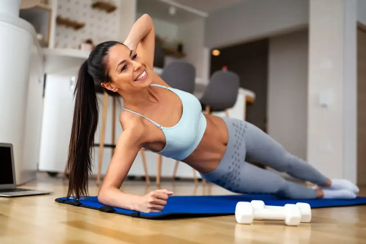 Mujer sonriente realiza una rutina de ejercicios en casa, haciendo plancha lateral sobre una esterilla azul. A su lado, pesas y un portátil.