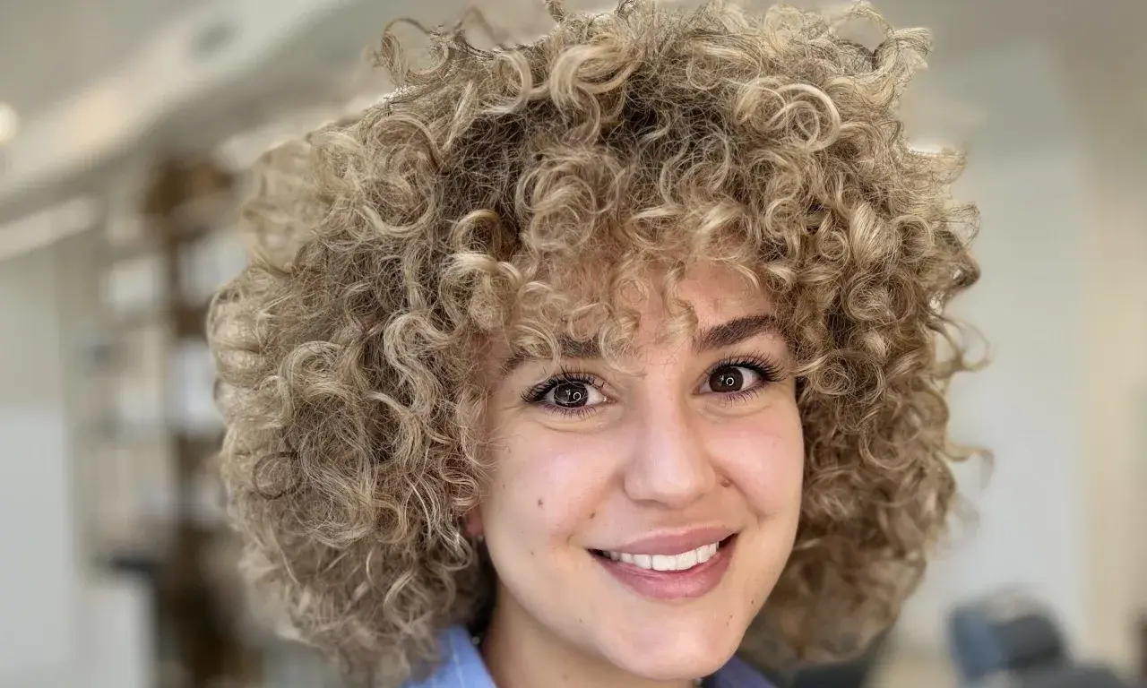 Sonrisa radiante de una mujer con un voluminoso flequillo pelo rizado rubio.