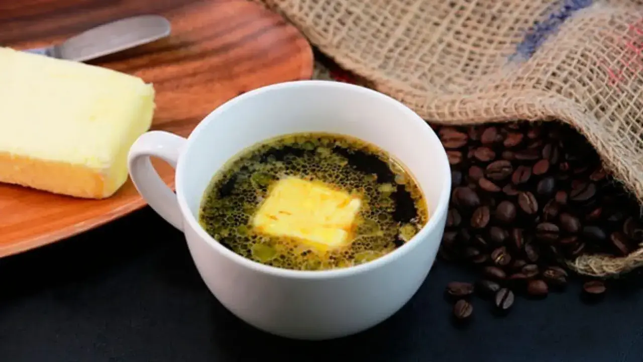 Una taza de café con mantequilla, junto a un trozo de mantequilla y granos de café tostados.