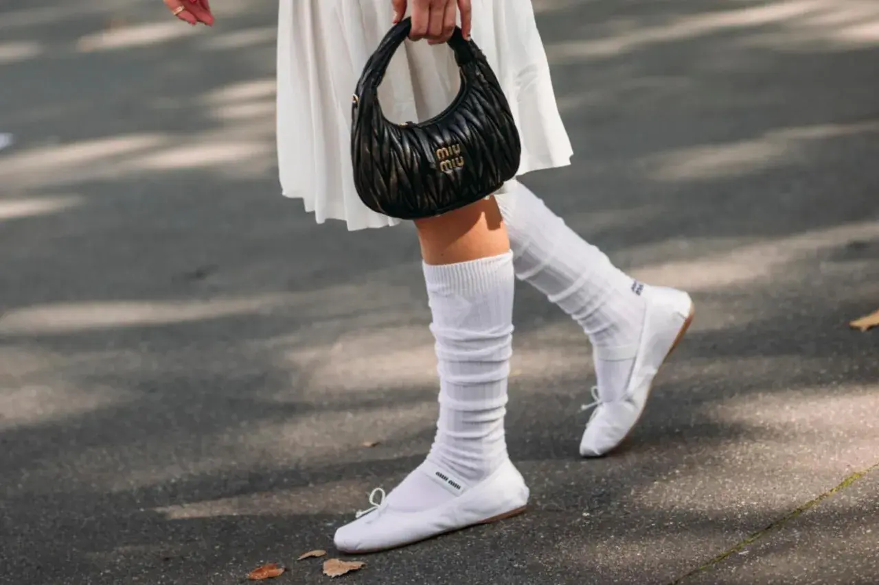 Piernas con bailarinas blancas y calcetines blancos arrugados, sosteniendo un bolso negro.
