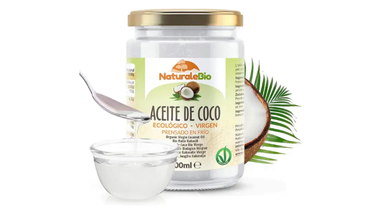 Un frasco de aceite de coco NaturaleBio con una cuchara vertiendo aceite en un cuenco. El aceite de coco es bueno para el pelo, nutriéndolo y dándole brillo.