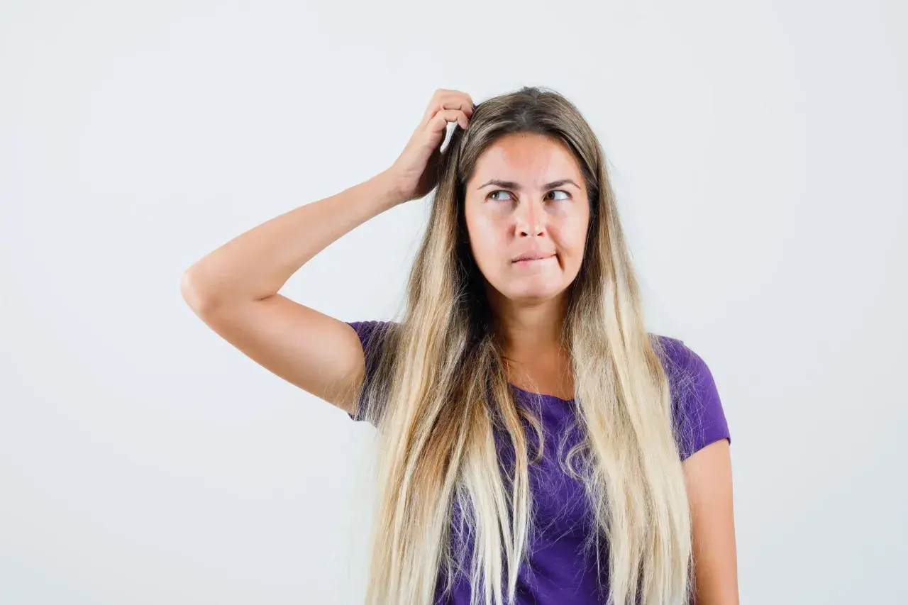 Mujer rubia con el pelo largo y suelto, se rasca la cabeza pensativa, buscando trucos de belleza para un look perfecto.