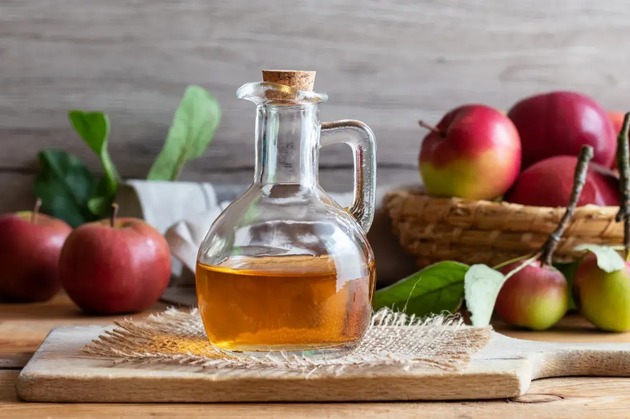 Botella de vidrio con vinagre de sidra de manzana, rodeada de manzanas rojas y verdes frescas.